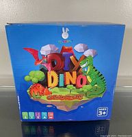 Front of a single box shows brand 'Squvbies', name 'DIY Dino Glow in the Dark', and age recommendation 3+. The box art includes colorful cartoon dinosaurs on a floating island and STEM/STEAM icons.