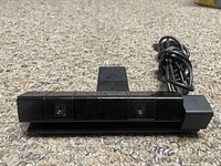 Front view of the PlayStation 4 camera webcam showing two camera lenses and cable on carpet background.