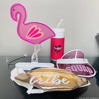 Pink flamingo light, travel mug, squad visor hat, and gold tribe fanny pack arranged on black tabletop.