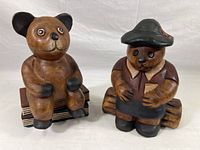 Front view of two hand painted teak wood bear figurines sitting on carved wooden logs, showcasing details of their faces and attire.