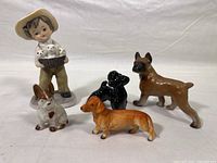 Photo showing five ceramic figurines together: one boy figurine holding a basket of white puppies, and four individual dog figurines of various breeds and colors