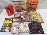 Photo showing full lot of 11 vintage cookbooks and recipe binders spread out, displaying colorful covers with food images and decorative art.