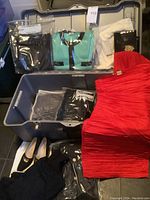 Overview of grey case containing Chanel sweaters, dust bags, clutch and red pleated dress with additional items on floor