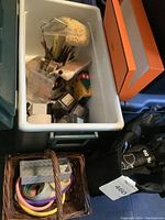 Green plastic container filled with various art supplies including makeup sponges, brushes, crayons, and rub-on transfers alongside pastel embroidery hoops in a wicker basket and a black soft cooler bag with blue trim.