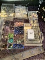 Clear plastic bead organizer with dozens of compartments filled with various semi-precious and glass beads plus tools visible at edge