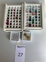 Two white trays of assorted faceted gemstones alongside two small boxes containing carved flower stones and emerald-cut stones