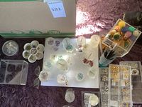 Top-down view of various gemstone beads in round plastic containers, pom poms, paint brushes and jewelry findings boxes on purple surface