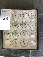 Box with 15 small circular compartments holding various small colorful cut gemstones including green, blue, pink, yellow, blackish, and red varieties.