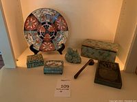 Various Asian decorative items on shelf including porcelain plate on stand, cloisonné boxes, celadon figurine, lacquered box, inkstone and wooden mallet