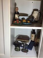 Two-shelf cabinet overview showing ceramic butler corkscrew, fruit plates, bowl, jug, bear pitcher, metal mixer attachment, books, whisky bottle, transistor radio