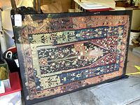 Full view of framed textile art piece leaning against objects in garage showing overall condition and pattern