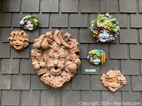 Six wall masks (three unglazed terracotta, two painted ceramic, one large terracotta grotesque head) mounted on shingle siding