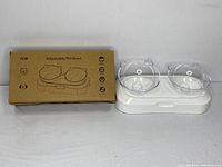 Photo of two clear plastic bowls with cat ear shapes on a white adjustable base with slide-out drawer beside the original cardboard box.