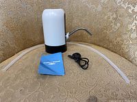 White automatic water dispenser placed on a patterned fabric surface with two translucent silicone tubes, a black USB cable, and an instruction manual.