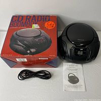 Photo showing the Proscan CD Radio Boombox, original retail box, black power cord, and user manual laid out flat on a white surface.