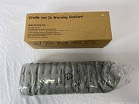 Heating pad wrapped in clear plastic beside its boxed packaging labeled 'Cradle you In Warming Comfort!', cardboard box providing product details and instructions.