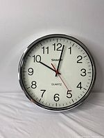 Front view of Sharp classic wall clock with white face, large black numbers, black hour and minute hands, red second hand, and silver frame.