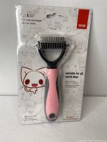 Front photo of the pet grooming brush in packaging; pink handle with black brush head and two rows of metal blades visible