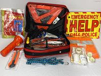 Photo showing open red carrying case with jumper cables, triangle warning signs, flashlight, emergency help sign, poncho, bungee cord, duct tape, and other emergency items.