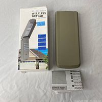 Wireless garage door keypad with flip-up cover and original packaging, shown with included instruction manual.