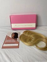 Box with pink packaging and instructional booklet alongside a blonde hair topper piece displaying its front and natural hair-like part.