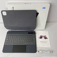 View of keyboard case open with visible keys, touchpad, and interior lining. Included user manual and charging cable in front beside product box.