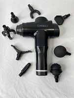 Black CARECJ handheld massage gun with all 8 different massage head attachments arranged around it on a white towel, showing various shapes and designs.