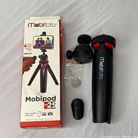 Complete Mobifoto Mobipod TT25 tripod set including box, tripod body, ball head, phone holder, and Bluetooth remote shutter button laid out on gray fabric surface.