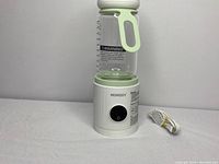 Liquid warmer base holding clear glass bottle with green handle attached, control button visible, charging cable next to it.