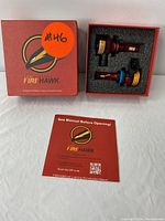 Two Firehawk LED light bulbs in red anodized metal housing placed in black foam tray inside a red box. Instruction manual with Firehawk logo and QR code below the box.