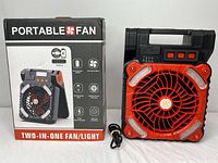 Front view of the 2-in-1 portable fan/light unit next to its retail box. Shows the device's black and orange design, including fan grill, LED light panels, handle, and red buttons on top. A USB Type-C cable included.