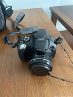 Canon PowerShot SX30 IS camera showing front angled view with lens cap on and strap attached on wood table.