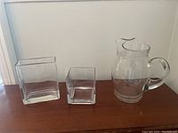 Photo showing the three clear glass items including one pitcher and two vases of different shapes and sizes on a wooden surface.