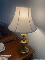 Brass table lamp illuminated with white bell-shaped fabric shade visible on wooden surface near a window and other items.