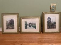 Three gold picture frames with matted photographs of French countryside scenes placed on wooden floor against grey wall under electrical outlet.
