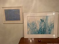Two framed prints placed on a wooden table and attached to the wall, one smaller Monet print with blue water lily scene and a larger nature print with tall grasses and a butterfly.