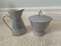 Photo showing a light grayish blue creamer and sugar dish with lid on cream carpet, items placed next to each other.