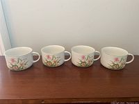 Four identical white ceramic soup mugs with floral patterns, shown from the front on a wooden surface.