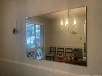 Wall mirror hanging above a ledge, reflecting a chandelier, chairs, windows with blinds and part of a dining room setup.