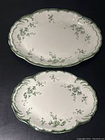 Two oval white ironstone platters with green flower decoration and scalloped edges viewed from above, showing full designs and sizes contrast.