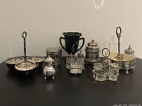 Wide shot showing assortment of antique silver and glass cruet sets and related items arranged on black surface against white background.