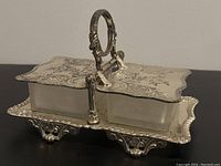 Front angle showing detailed engravings on hinged lids and silver plated handle above two glass inserts.