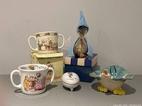 Photo of lot with two Bunnykins mugs, Winnie the Pooh mug, Holly Hobby trinket box, silver plated duck piggy bank wearing blue knit hat and sweater, and ceramic duck bowl.