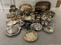 Photo showing a wide view of the large grouping of various silver plated serving pieces arranged on the floor. Items include serving trays, dishes, bowls, candle holders, and utensils, some with intricate floral and leaf embossing.