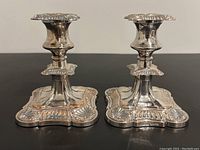 Front view of both ornate silver plated candle holders side by side on a black surface
