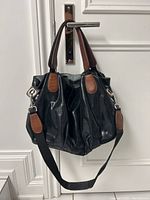 Black textured leather handbag hanging on a door handle showing entire bag and detachable shoulder strap with brown leather handles and accents.