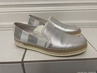Side view showing shiny silver striped leather upper, elastic side accents, and natural jute braided trim over white rubber sole.