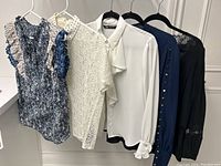 Five women's blouses hung on hangers showing front view including different colors and details like ruffles, lace, buttons.