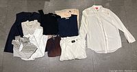 Lot of folded women's shirts, sweaters and tops including white button-up shirt on hanger, striped and brown tops, white eyelet blouse, and more