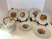 Six bareuther dessert plates with gold rims and various fruit images displayed on stands behind a white porcelain teapot with bamboo handle.